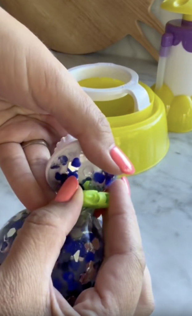 Close-up of hands holding a broken stress ball with blue and silver confetti inside; a yellow juicer and purple bottle are visible in the background on a marble counter.