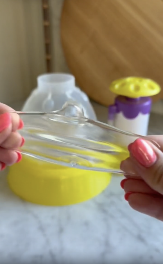 Hands with pink nails stretch clear slime in front of a yellow and purple toy and a large wooden board on a marble surface.
