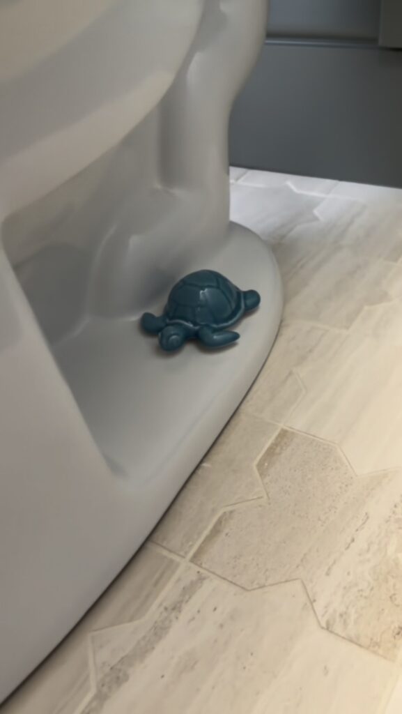A small blue turtle-shaped object rests at the base of a gray toilet on a light-colored, patterned tile floor.