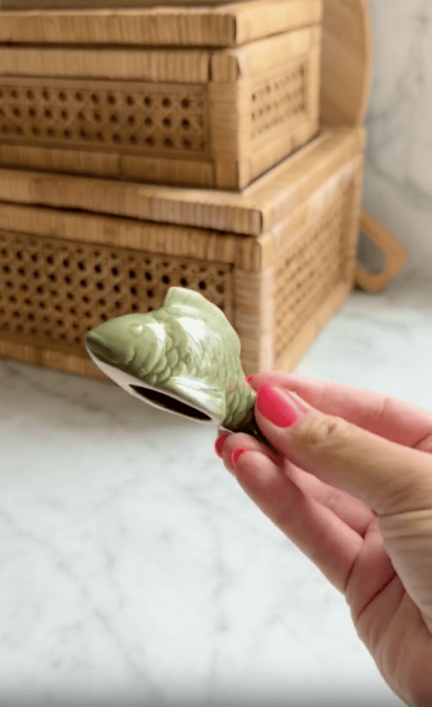 A hand with pink nail polish holds a small, green, fish-shaped ceramic object in a kitchen with woven wooden containers in the background.