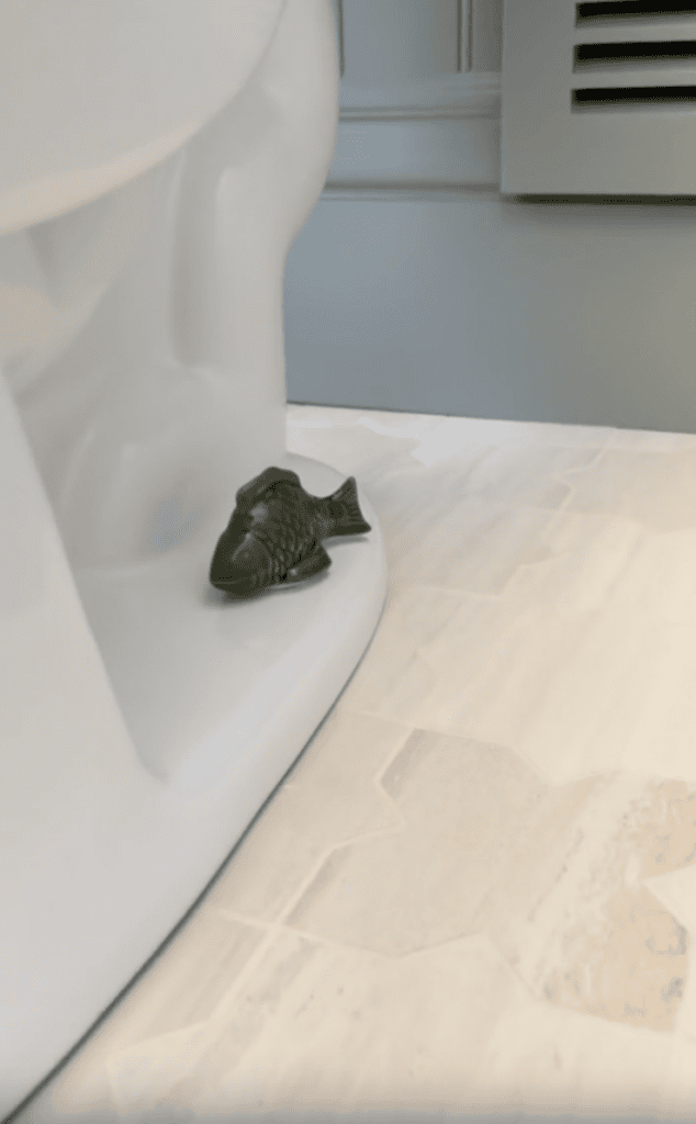 A small green fish figurine is placed at the base of a white toilet on a beige-tiled bathroom floor near a vent.