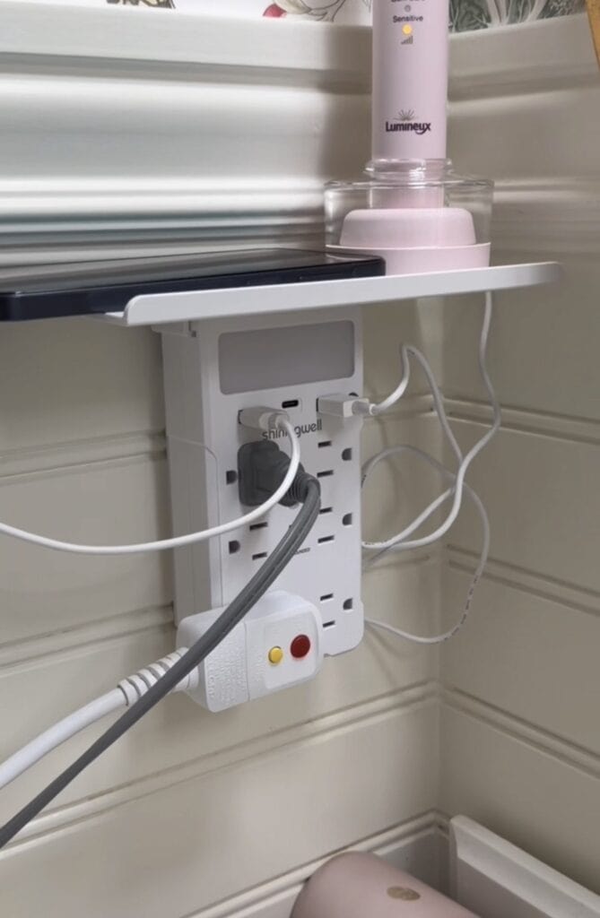 A wall outlet extender with multiple plugs and USB charging cables is mounted under a small white shelf. A bottle and a phone rest on the shelf. The background is white paneling.