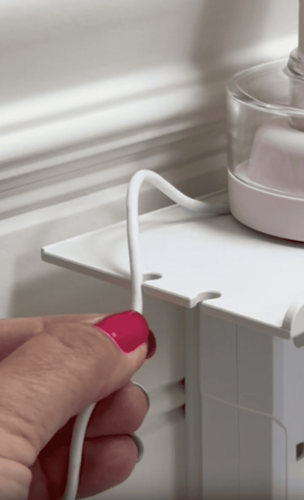A hand with red nail polish threads a white cord through a notch on a white plastic organizer shelf beside a pink device.