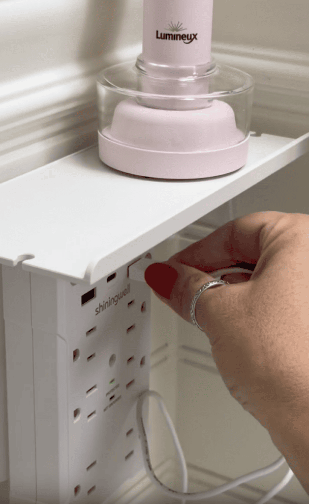 A hand with red nail polish plugs a cord into a white wall outlet extender beneath a shelf holding a pink Lumineux container.
