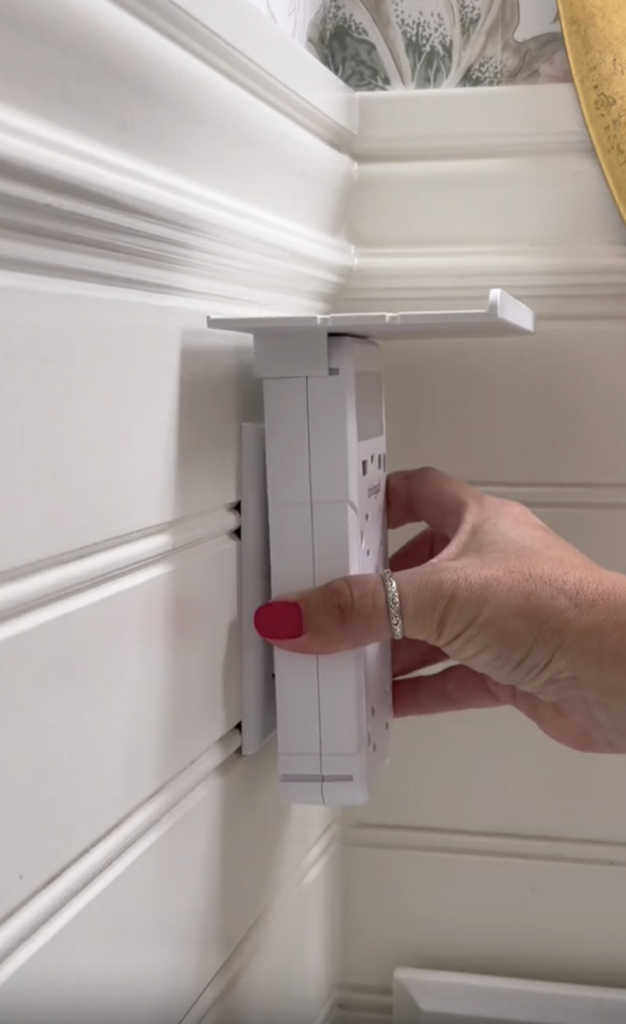A hand with pink nails and a silver ring pulls open a hidden white wall panel, revealing a secret compartment behind it.