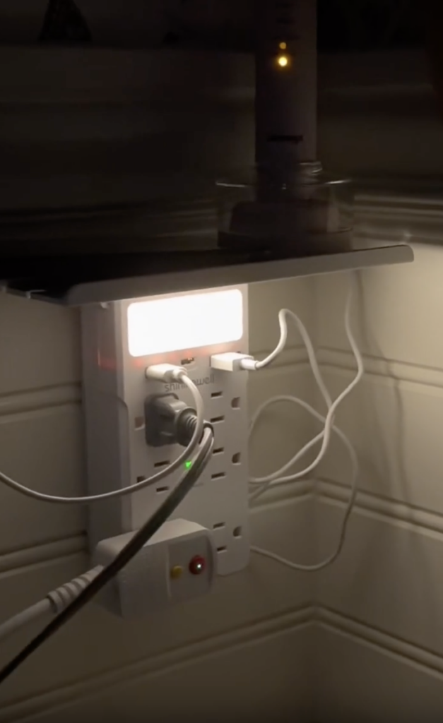A wall outlet extender with multiple plugged-in devices and a built-in night light glows in a dimly lit room, with several cords plugged into different sockets.
