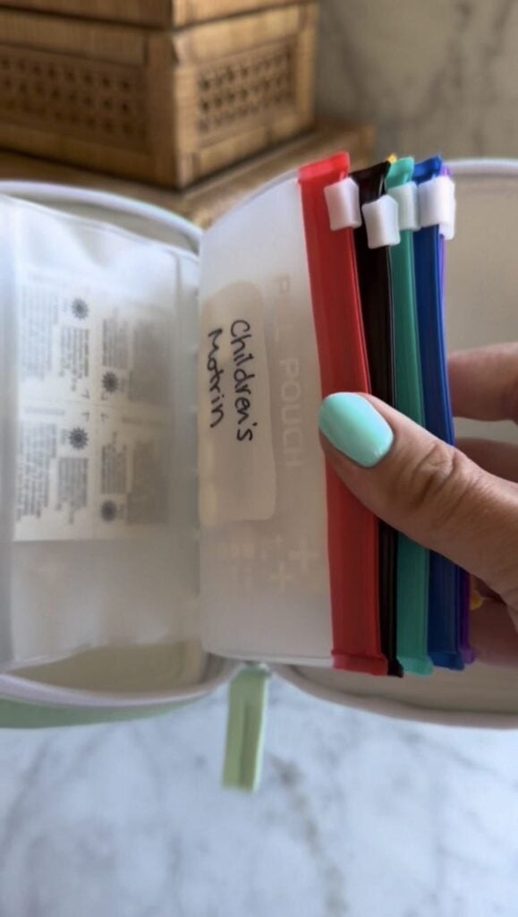 A hand with turquoise nail polish holds color-coded zip pouches in a first aid kit. One pouch is labeled “Children’s Motrin.” The background shows a marble surface and a wicker box.