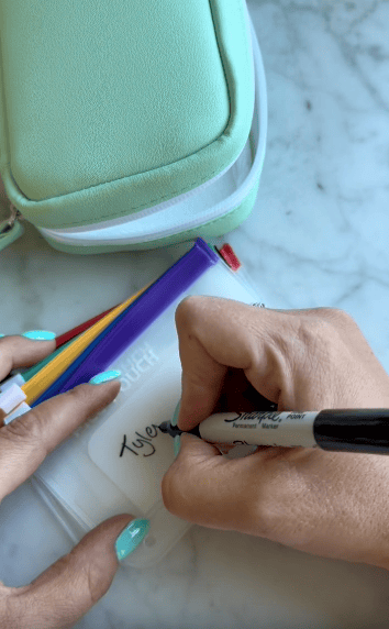 A person with teal nails writes “Tyler” on a clear pouch with a black Sharpie marker. Colored pens are inside the pouch, and a mint green bag is nearby on a marble surface.