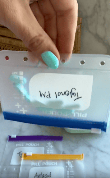 A hand with turquoise nail polish holds a clear pill pouch labeled “Tylenol PM” upside down. Two other labeled pill pouches are visible below on a marble surface.