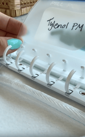 A hand with turquoise nail polish holds an open binder labeled “Tylenol PM,” displaying plastic pockets with rings for organizing pills.