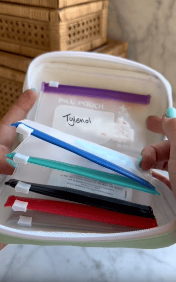 A hand holds open a pouch containing several labeled, colored pill pouches; the top pouch is labeled “Tylenol” in black marker.