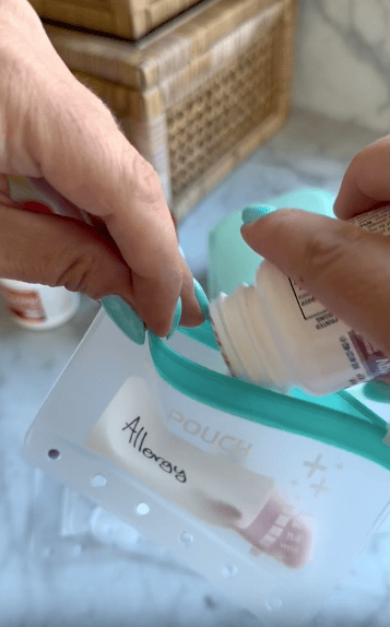 A person pours pills from a bottle into a zippered pouch labeled “Allergy.” The person’s fingernails are painted teal. Basket and bottle are blurred in the background.