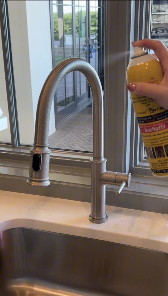 A hand sprays cleaner from a yellow can onto a silver kitchen faucet above a sink, with a window and patio visible in the background.