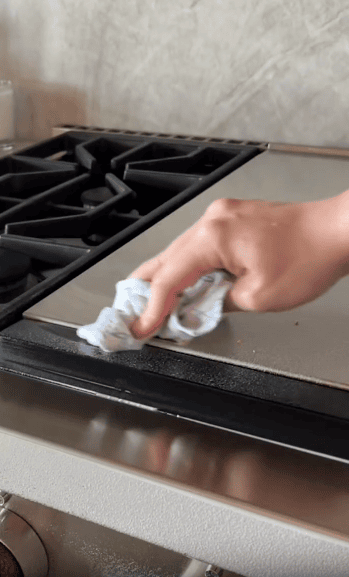 A hand uses a cloth to wipe the edge of a stovetop, cleaning near the burners.