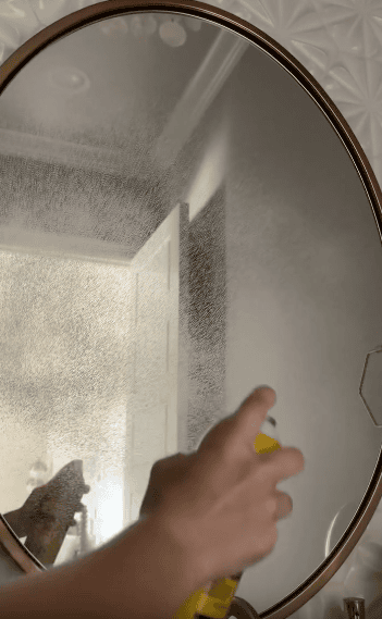 A hand sprays cleaner onto a round bathroom mirror, creating a misty pattern on the glass. The background shows a partially open white door and patterned white wall tiles.