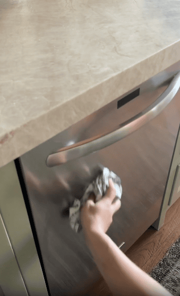 A person wipes the stainless steel front of a dishwasher with a cloth, cleaning the surface beneath a countertop in a kitchen.