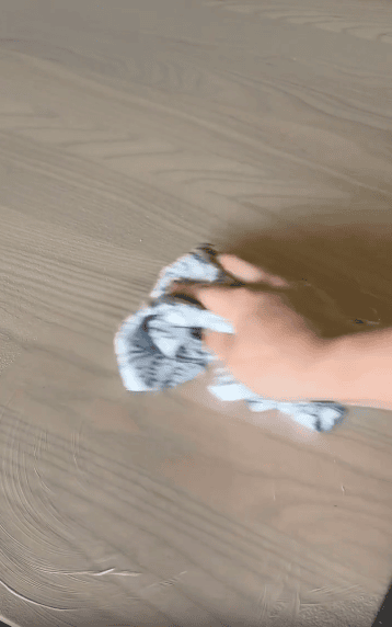 A hand wipes a light wooden surface with a gray and white cloth.