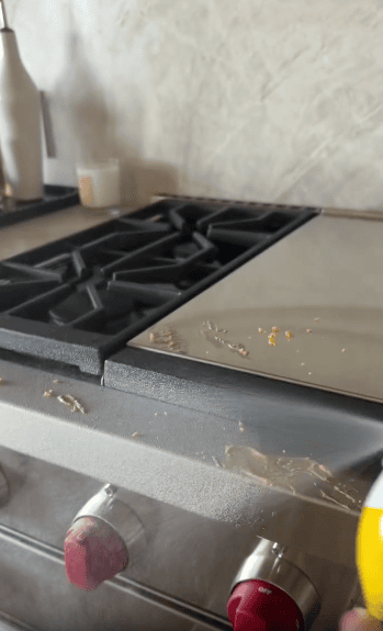 A person sprays cleaning product onto a stainless steel stovetop with black grates and red knobs, preparing to clean food residue.