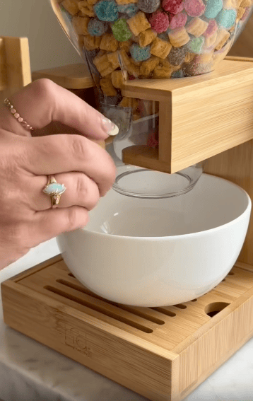 A hand turns the knob on a wooden cereal dispenser, dropping colorful cereal into a white bowl below.