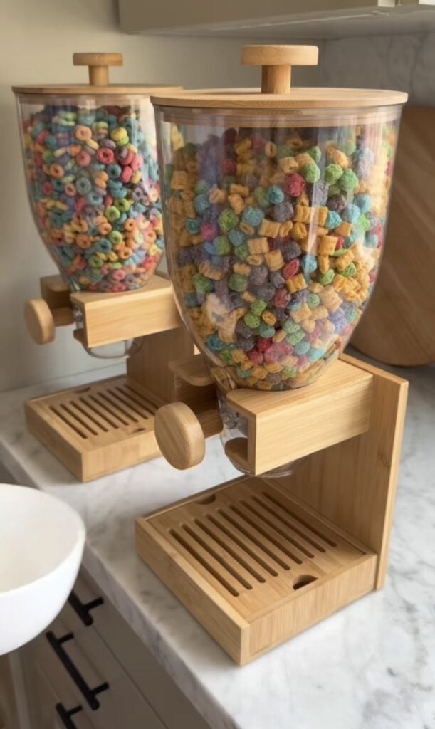 Two wooden cereal dispensers on a marble countertop, one filled with colorful round cereal and the other with multicolored square cereal, with a white bowl placed next to them.