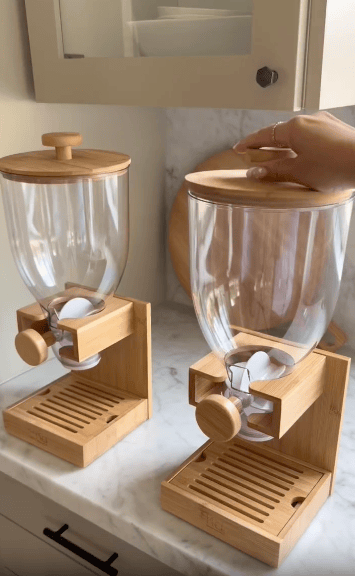 Two clear and bamboo cereal dispensers sit on a marble counter. A hand lifts the wooden lid of one dispenser. The background includes white cabinetry and dishes.