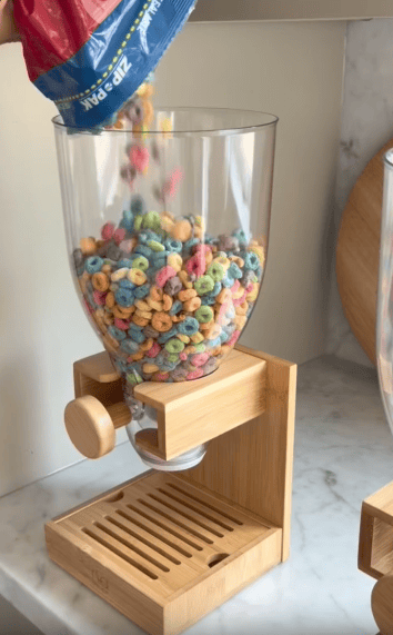 A hand pours colorful cereal loops from a bag into a clear dispenser with a wooden base, set on a marble countertop.