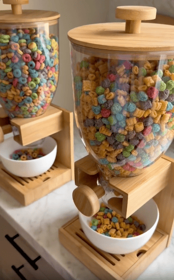 Two wooden cereal dispensers filled with colorful cereals, each dispensing into a white bowl below, sit on a marble countertop.