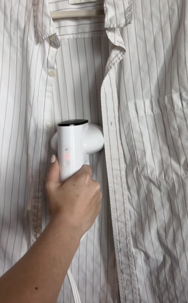 A hand holds a white handheld steamer, removing wrinkles from a light-colored, striped button-up shirt hanging on a rack.