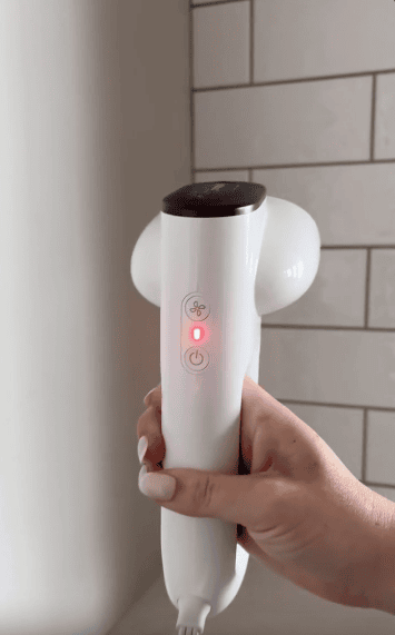 A hand holds a white handheld device with a red power indicator light, in front of a tiled wall.