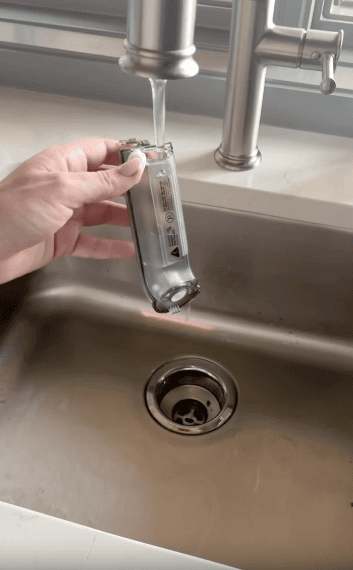 A hand holds an electric hair clipper under running water from a kitchen faucet, rinsing it above a stainless steel sink.