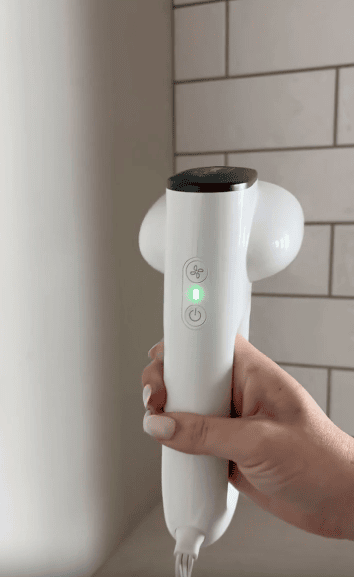 A hand with light-colored nail polish holds a white handheld device with two buttons and a glowing green light, in front of a tiled wall.