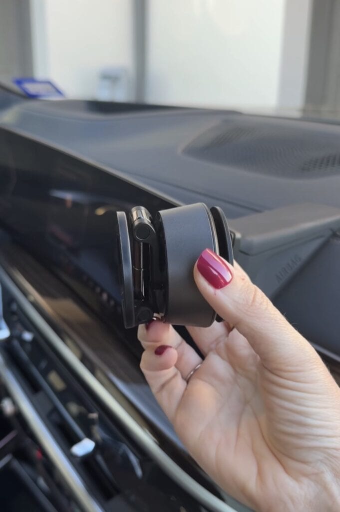 A person with red nail polish holds a black magnetic phone mount inside a car, positioned near the dashboard and air vent.