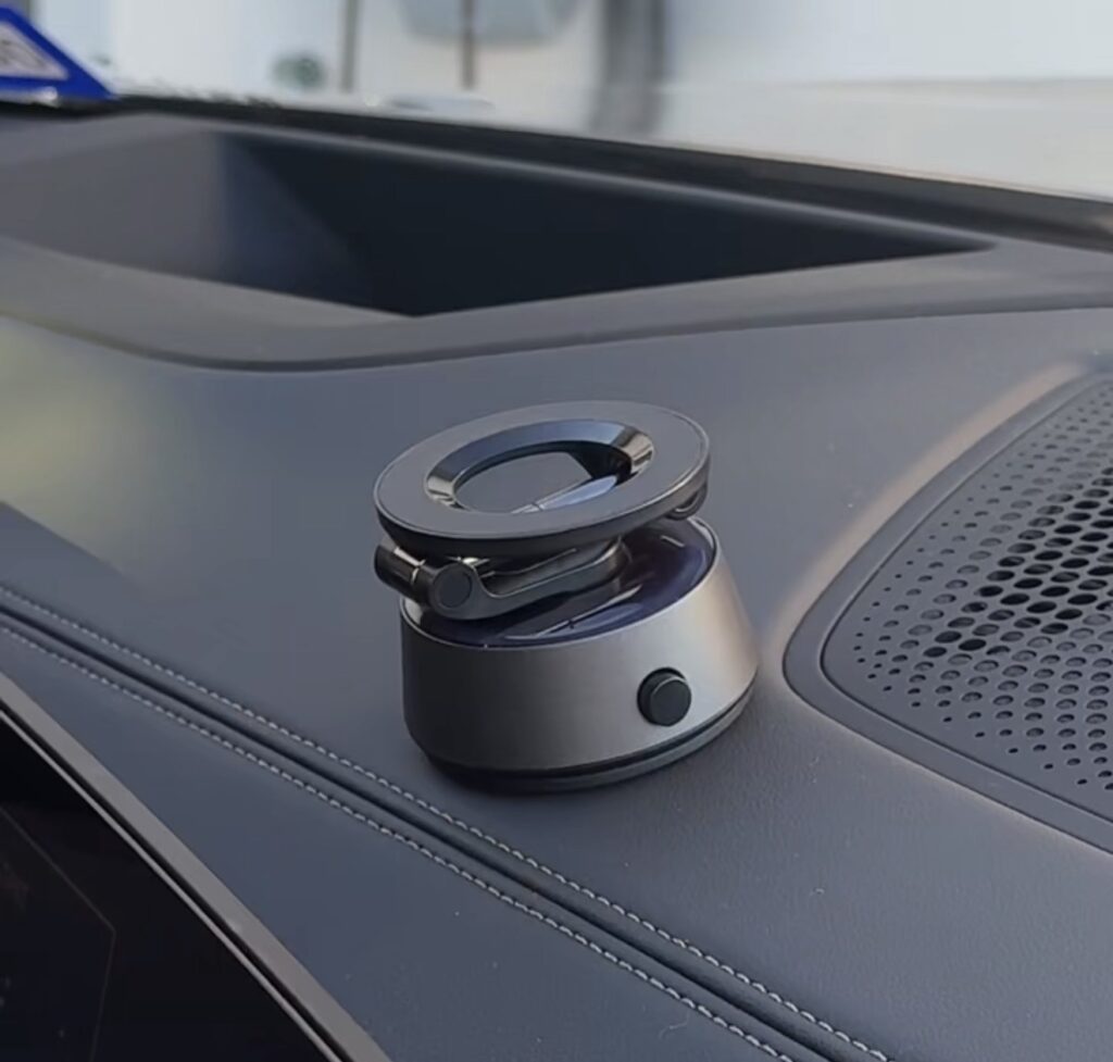 A small, round, metallic device with a circular top sits on a car dashboard near an air vent, featuring a button on its side.