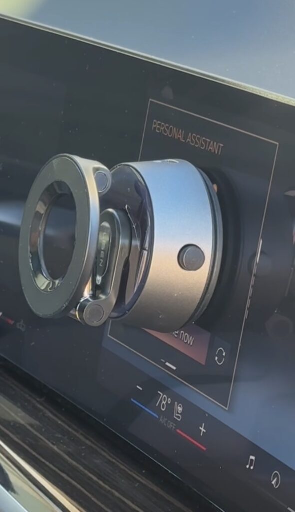 A close-up of a metallic car mount attached to a touchscreen display, which shows the text “PERSONAL ASSISTANT” and climate control settings at the bottom.