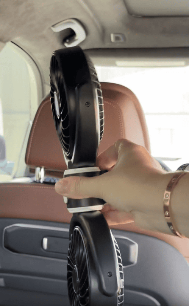 A hand holds a double-headed black fan inside a car with brown leather seats. The fan is positioned vertically, and sunlight is coming through the car window.