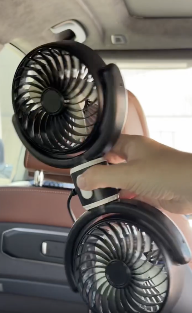 A hand is holding a small, portable dual-blade fan inside a car with brown leather seats and a window in the background.