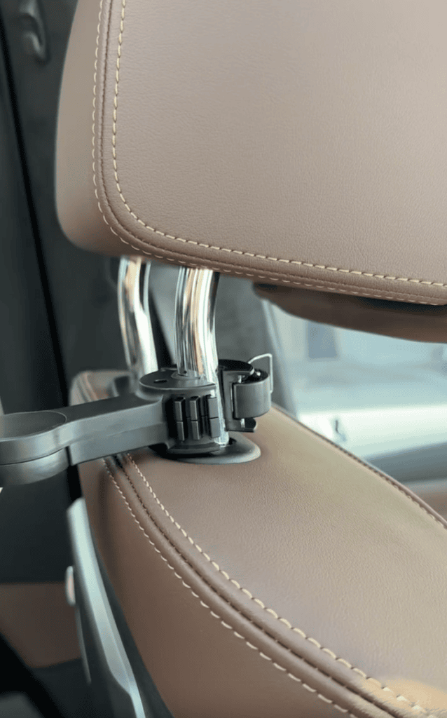 A close-up of a car seat headrest with a black plastic mount attached to the metal rods, possibly for holding a device or accessory. The seat is brown leather.