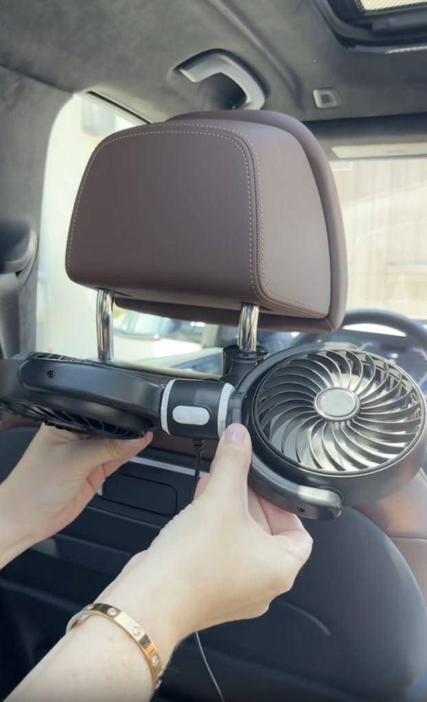 A person attaches a small, dual-blade fan to the back of a car seat headrest. The fan is black and powered by a cord.