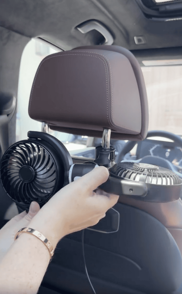 A person installs a double fan onto the back of a car seat headrest, securing it for rear passengers.