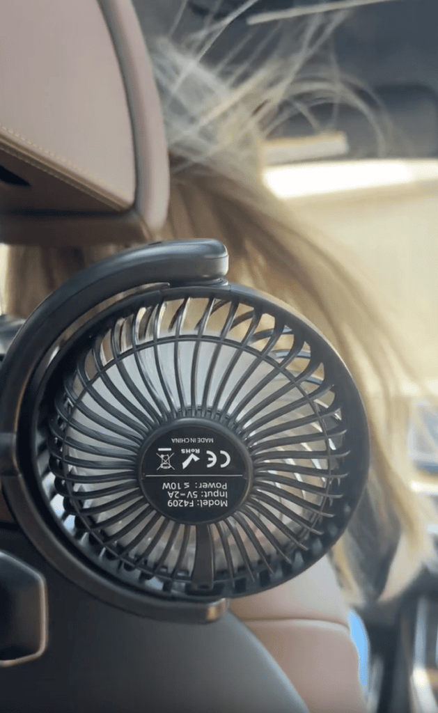 Close-up of a small black fan attached to a car seat headrest, with a person’s long hair blowing in the breeze behind it. Sunlight streams in through the car window.