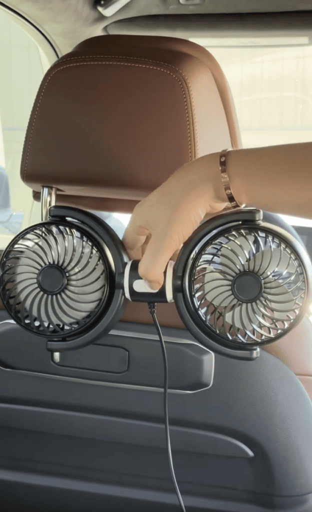 A hand adjusts a dual-blade fan attached to the back of a brown car seat headrest, with a charging cable plugged in.