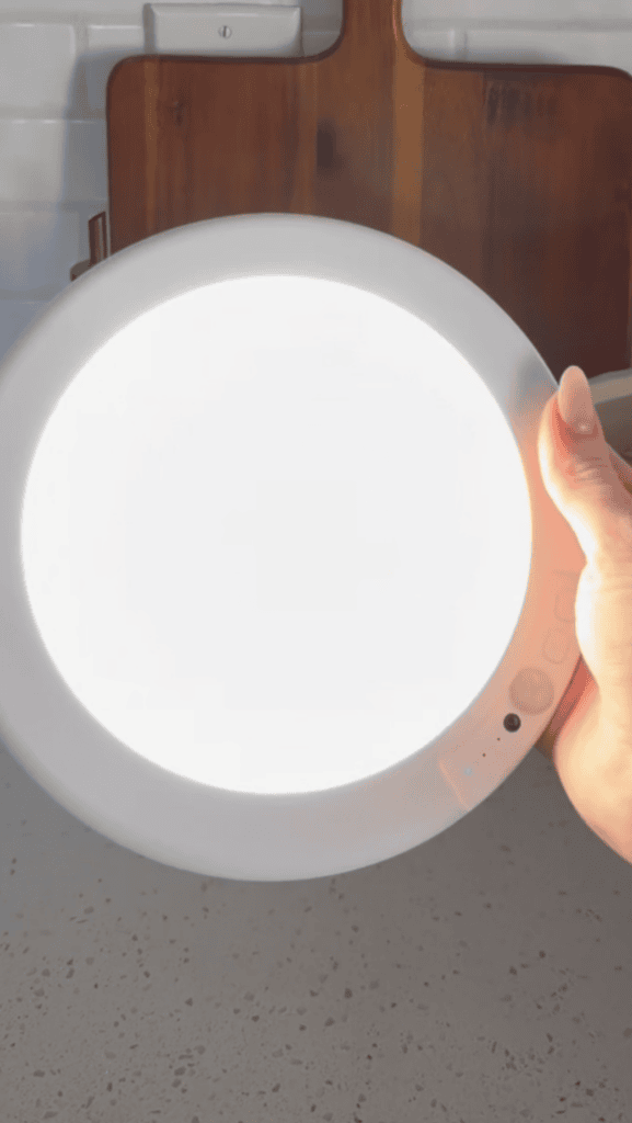 A hand holds a bright, round LED light panel above a speckled countertop, with a wooden board and white tile backsplash in the background.