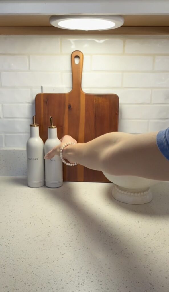 A hand reaches for a white oil dispenser beside a matching vinegar bottle on a kitchen counter, with a wooden cutting board and white tiled backsplash in the background.