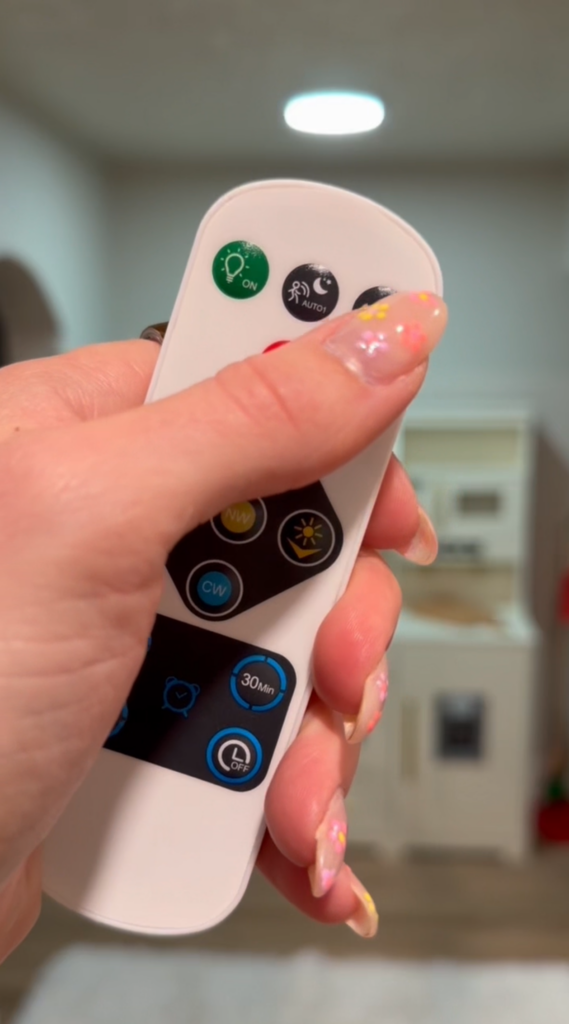 A hand with pastel-painted nails holds a white remote control with various black, blue, and green buttons, including one with a lightbulb icon. A softly blurred kitchen is visible in the background.