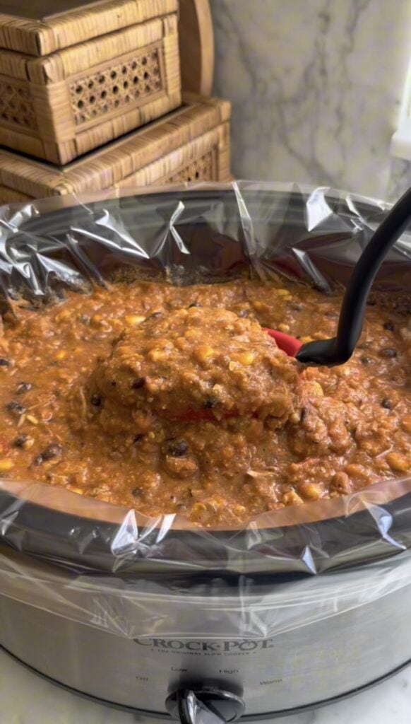 A slow cooker filled with chunky chili containing beans, corn, and tomatoes. A ladle is lifting a serving of the chili, and the slow cooker is lined with a clear plastic liner. Wicker baskets are in the background.