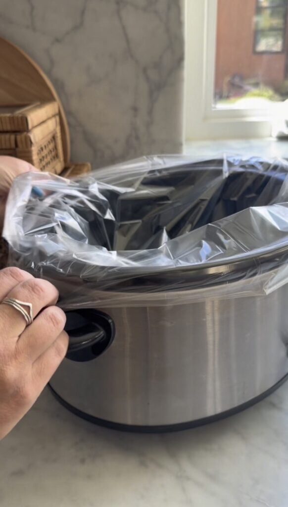 A hand pulls a clear plastic liner over the edge of a stainless steel slow cooker on a kitchen counter with a marble backsplash.