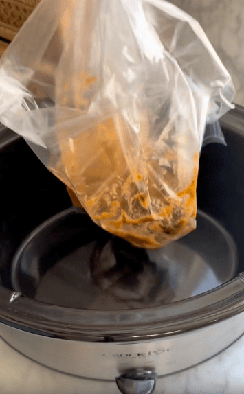 A plastic bag containing marinated food is being placed into a black Crock-Pot slow cooker on a marble countertop.