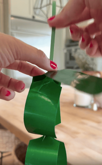 A person with red nail polish holds and curls a green ribbon, preparing it for decoration on a wooden kitchen countertop.