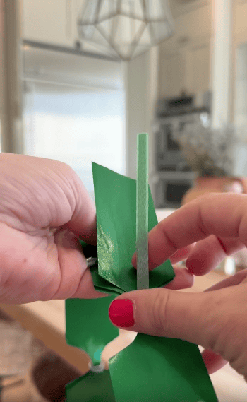 Close-up of a person with pink nail polish folding shiny green ribbon, with a light-filled kitchen in the background.