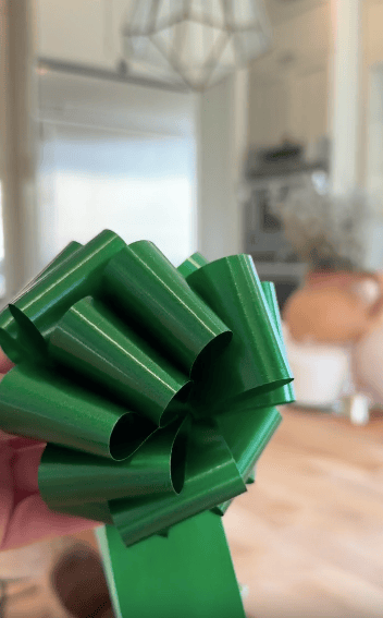 A hand holds a shiny green gift bow indoors, with a modern kitchen and blurred background visible.